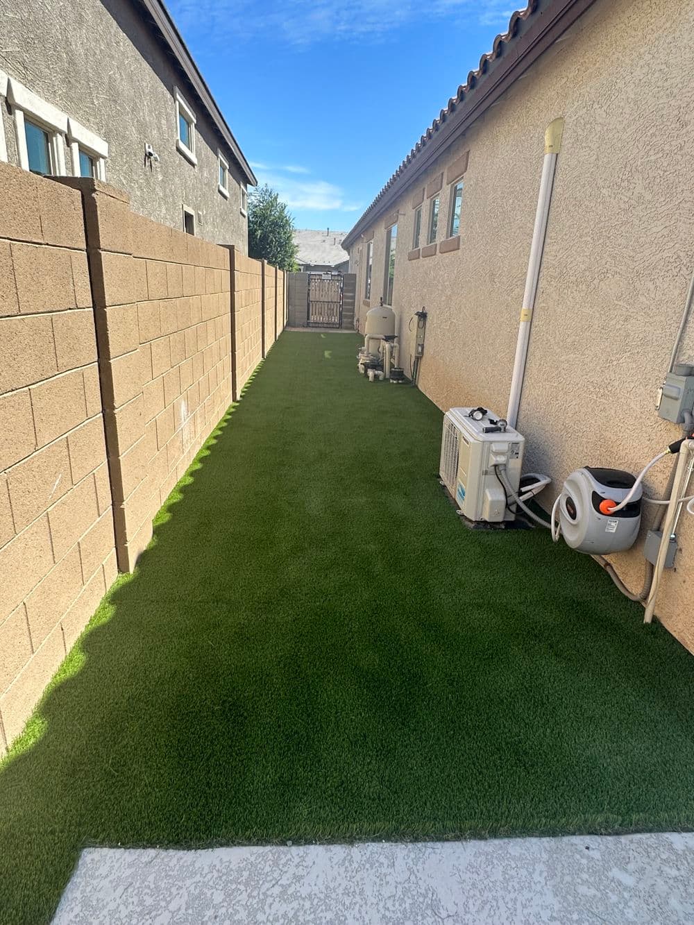 Narrow side yard with artificial turf, walls, and utility equipment under a clear blue sky.