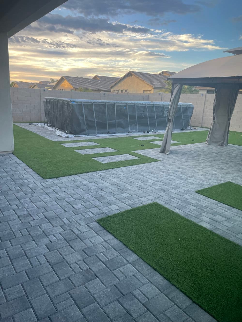 Backyard with paver stones, artificial grass, and a covered above-ground pool at sunset.