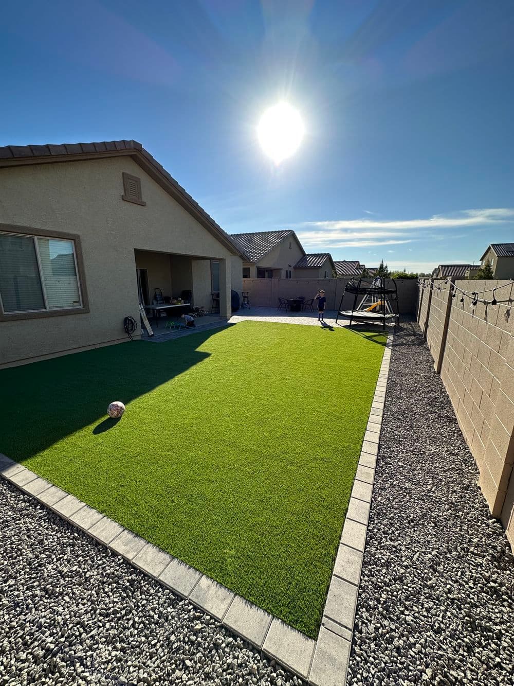 Sunlit backyard with artificial grass, a soccer ball, and a wall with houses in the background.