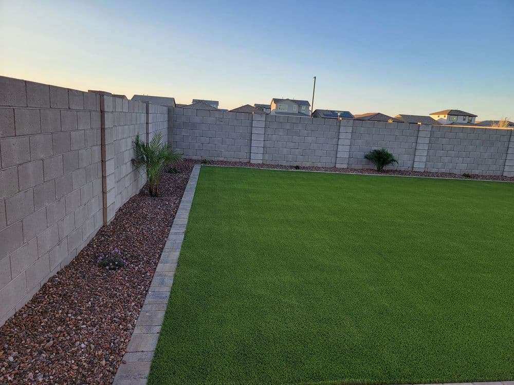 Lush green lawn bordered by stone path and gray block wall in suburban backyard setting.