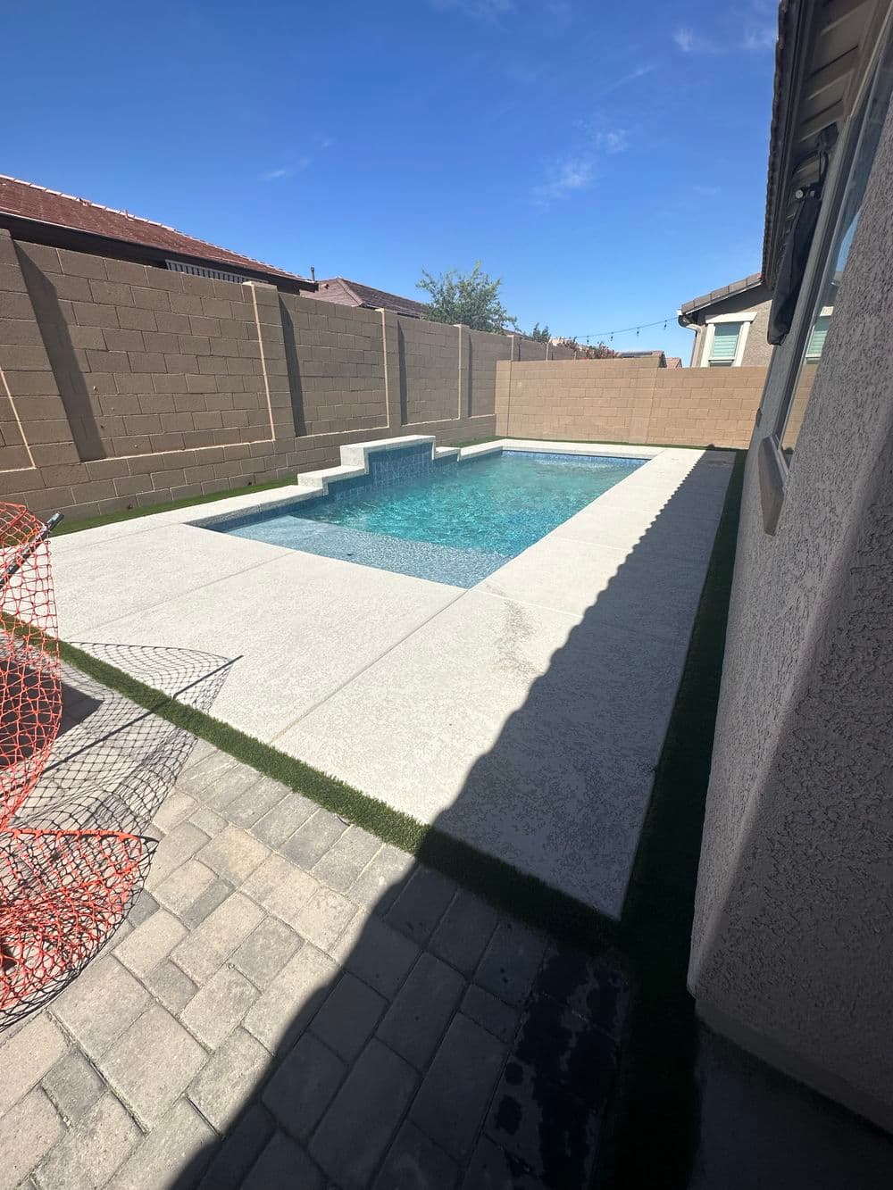 Modern backyard pool with concrete deck, surrounded by a block wall and clear blue sky.