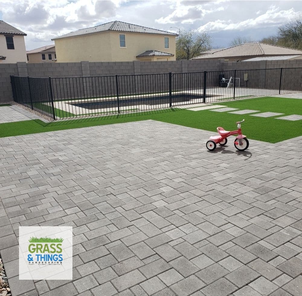 Paved backyard with artificial grass, black fence, and a red tricycle on the patio.