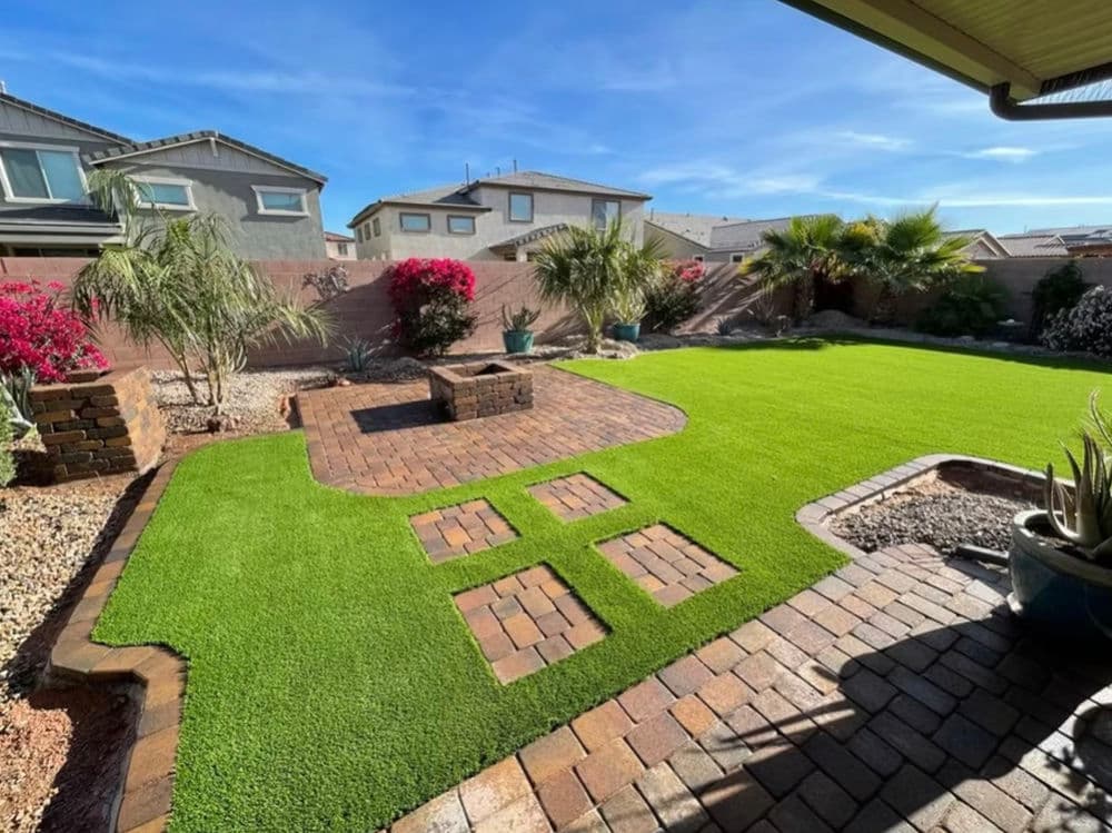 Lush backyard with synthetic grass, stone patio, and colorful landscaping in sunny setting.