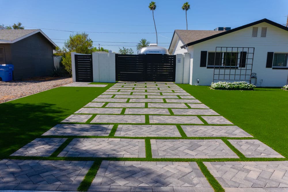 Modern driveway design with gray pavers and artificial grass, surrounded by palm trees.