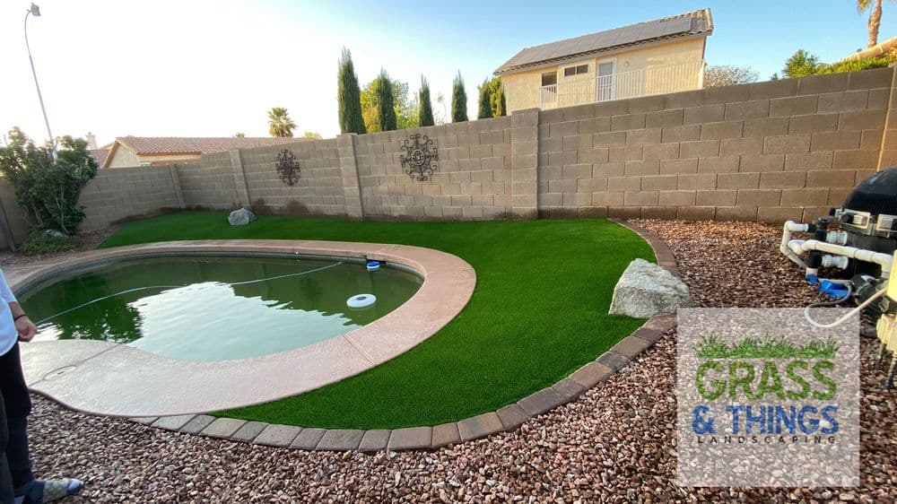 Lush artificial grass around a swimming pool with stone landscaping and a residential backdrop.