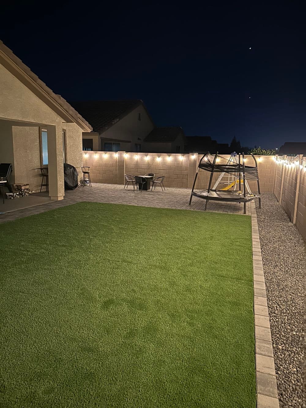 Backyard at night with grass, swing set, and string lights illuminating the space.