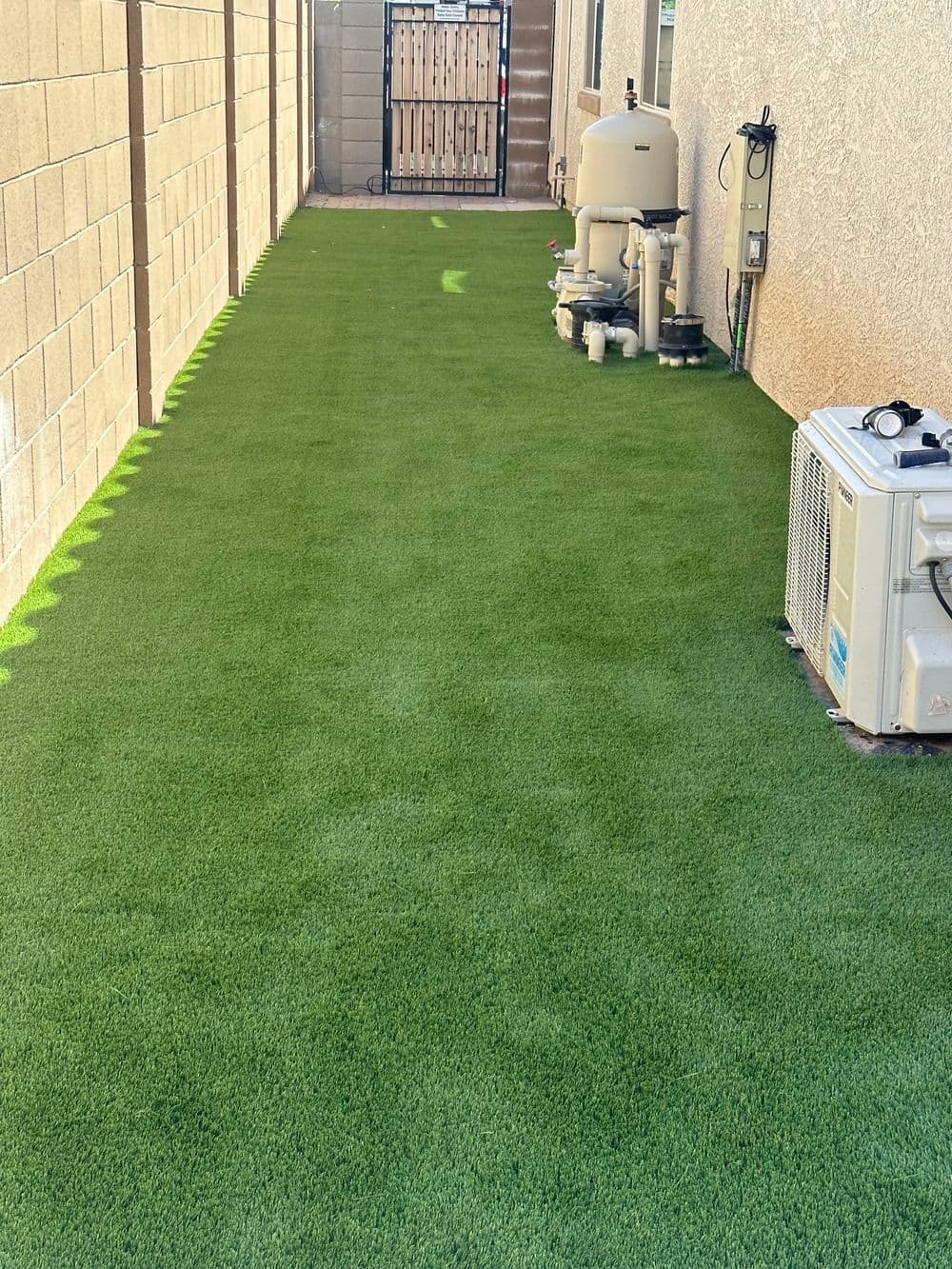 Lush green artificial turf in a narrow backyard space with utility equipment nearby.