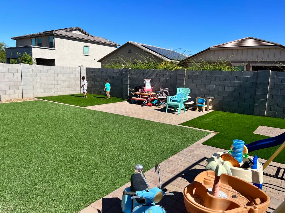Children playing in a backyard with artificial grass, patio area, and colorful toys.