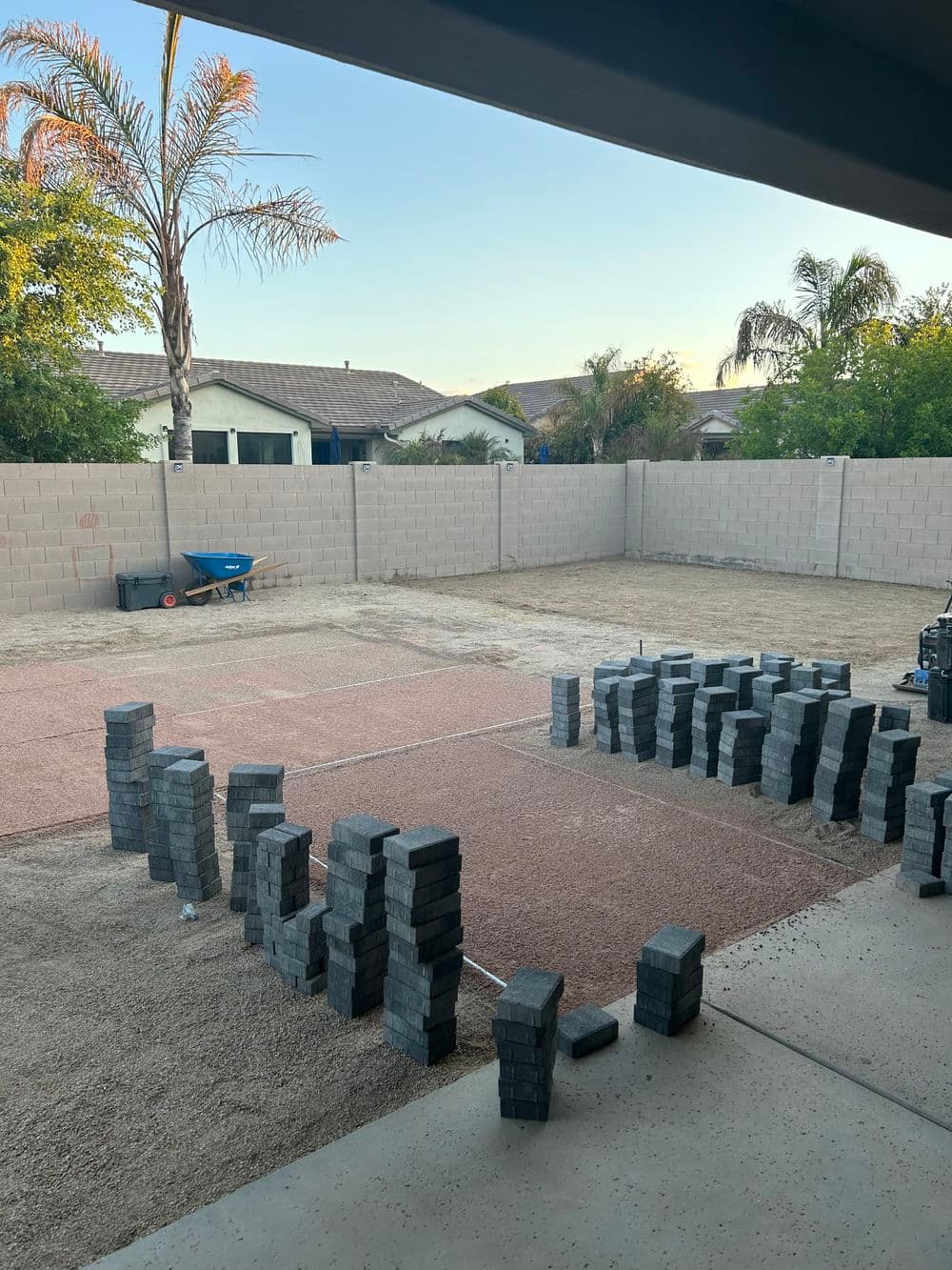 Paved backyard project with stacked gray bricks and a wheelbarrow against a clear sky.