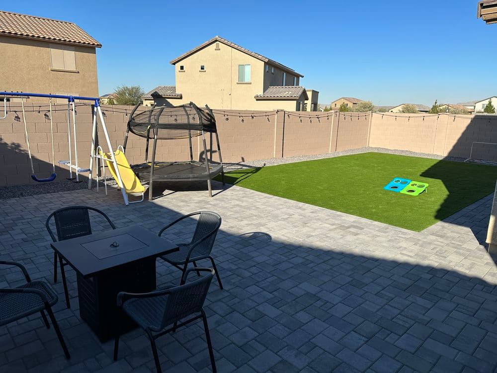 Backyard with synthetic grass, trampoline, swing set, and patio furniture under clear blue sky.